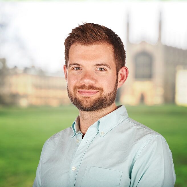 A man with a beard and short brown hair is smiling while wearing a light blue button-up shirt. He stands outdoors with a blurred background of grass and a large, historic building. The sky is clear.