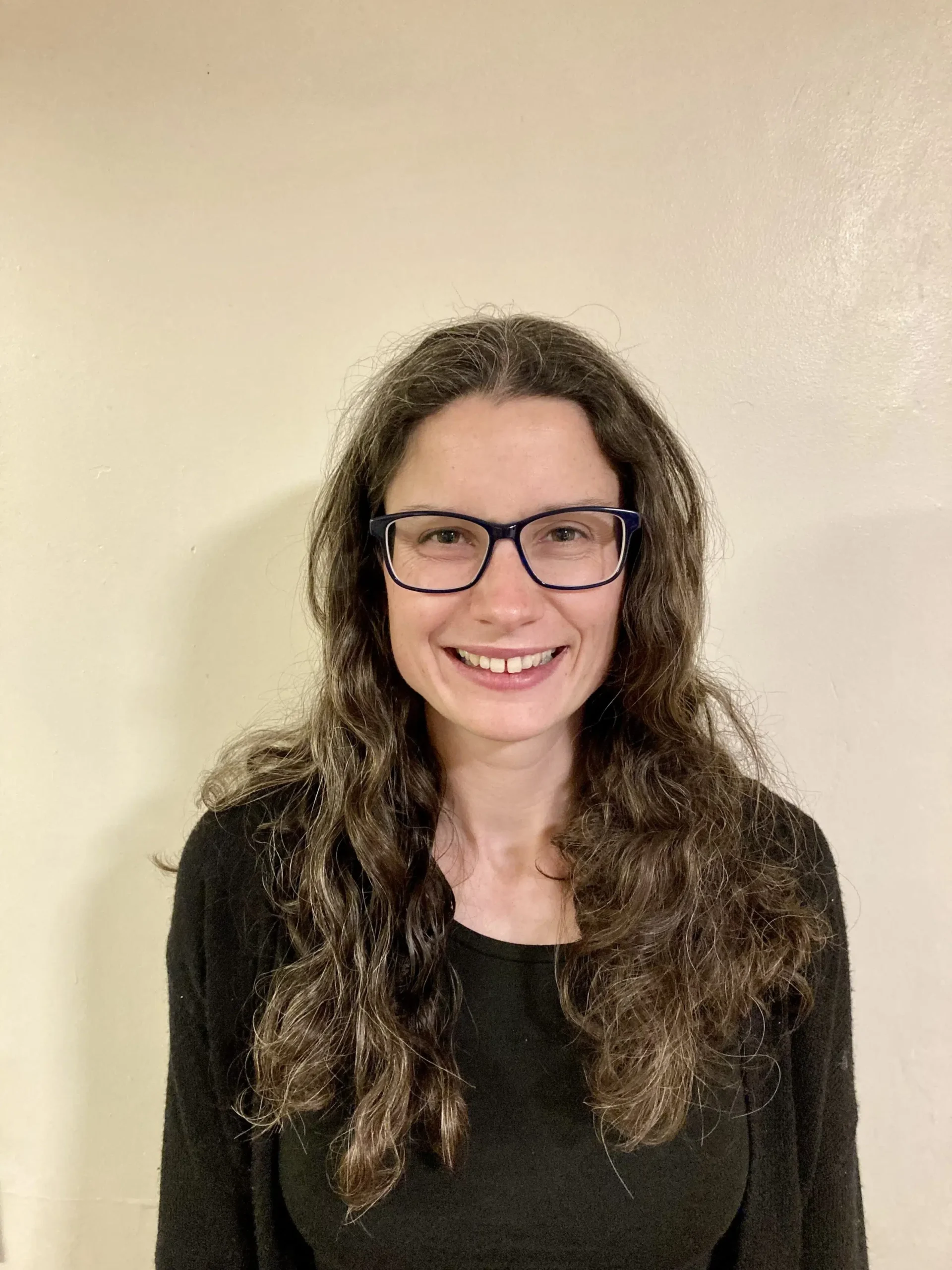A woman with long, wavy brown hair and glasses smiles at the camera. She is wearing a black top and standing against a plain beige wall.