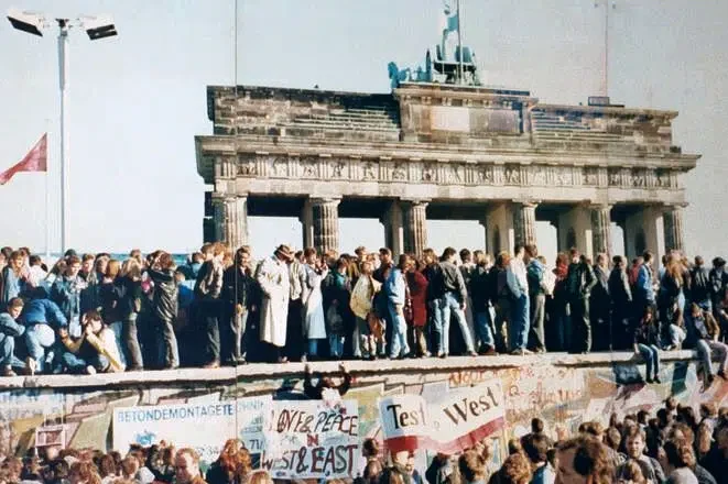 Crowds gather around the Berlin Wall near the Brandenburg Gate during its fall in 1989, marking a historic moment of unity and freedom. The wall, adorned with graffiti and banners, stands as a vibrant testament to the power of politics in reshaping history.