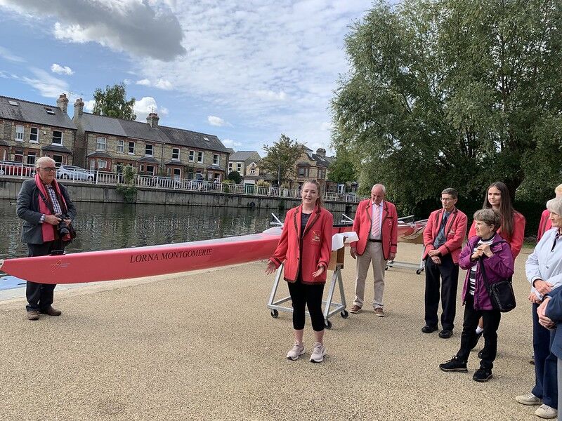 A woman in a red jacket stands smiling beside a red rowing scull named Lorna Montgomerie on a riverside promenade. She is surrounded by people, some also wearing red jackets. In the background, there are houses and trees.