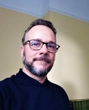 Man with short hair, glasses, and a beard, wearing a black sweater, stands in a room with yellow walls and a bookshelf in the background.