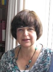 A person with short dark hair is wearing a patterned top and a necklace with green beads. They are standing in front of a window with light curtains and a bookshelf.