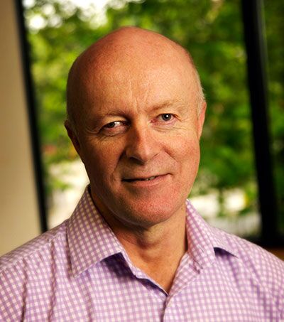 A bald man wearing a purple checkered shirt is smiling at the camera. He is indoors, standing in front of a window with a view of green foliage outside.