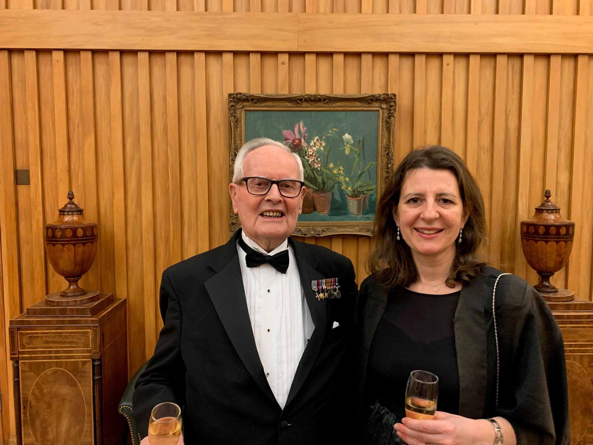 An elderly man in a tuxedo with medals and a woman in a black outfit stand smiling, each holding a glass of champagne. They are in a wood-paneled room with ornate vases.