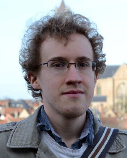 A person with curly hair and glasses is wearing a light-colored jacket over a sweater and shirt.