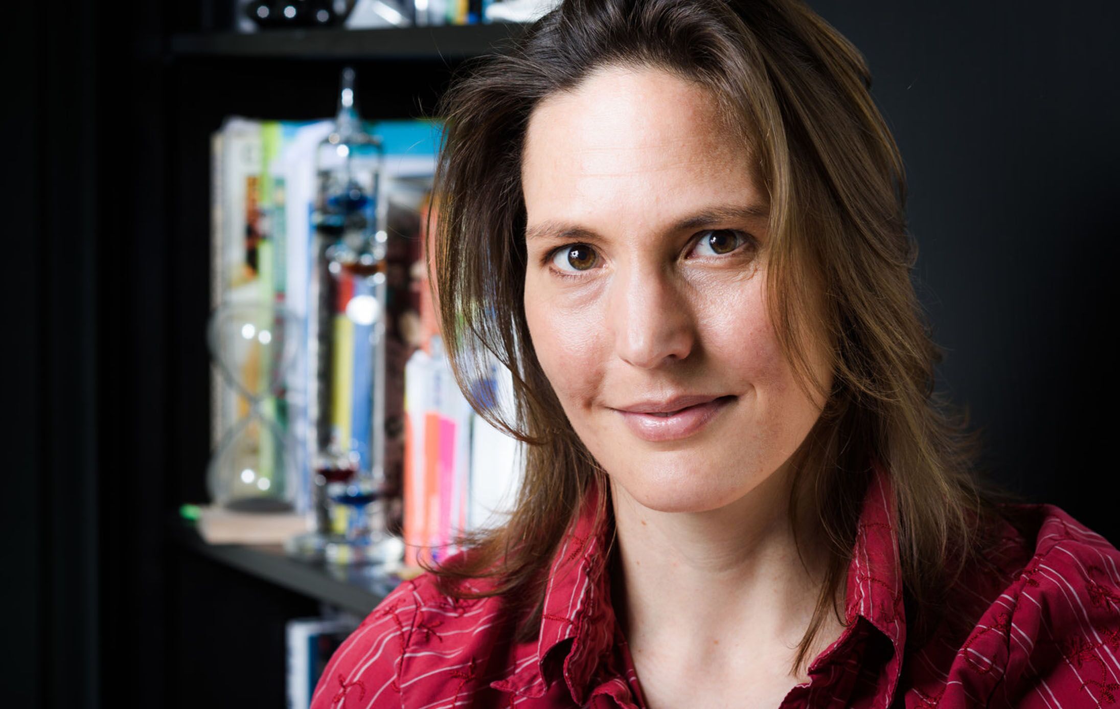 A person with shoulder-length brown hair, wearing a red shirt, smiles gently while seated in front of a bookshelf filled with books and a decorative hourglass.