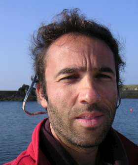 A man with short, dark hair and facial stubble is outdoors near a body of water. He is wearing a red jacket and has sunglasses hanging around his neck. The background shows a clear blue sky and a distant shoreline.