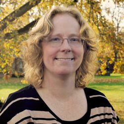 A woman with curly blonde hair and glasses smiles while standing outdoors. She is wearing a black and tan striped top. The background features trees with autumn leaves and a grassy area.