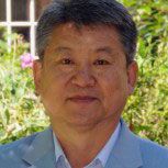 A person with short gray hair wearing a light blue blazer is looking at the camera. The background features greenery and a partial view of a building with a window.