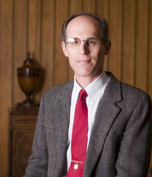 A man with glasses, wearing a gray blazer, white shirt, and red tie, is sitting in front of a wooden wall. Theres a decorative urn on a small cabinet behind him.