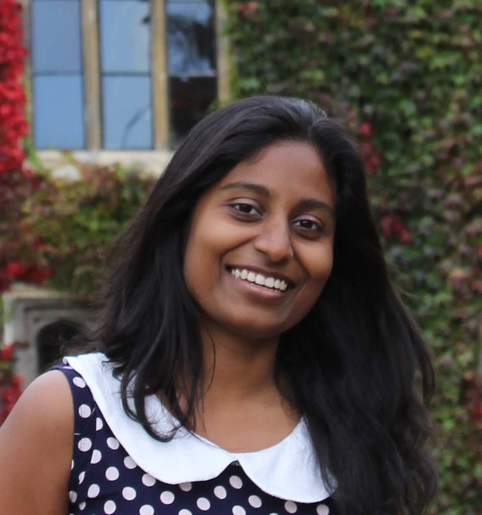 Smiling woman with long dark hair, wearing a polka dot dress with a white collar, stands outside. There's a vine-covered wall and a window in the background.