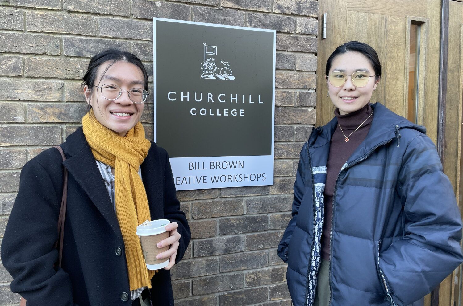 Two students stand together for a photo in front of the Churchill College sign, representing their connection to the institution
