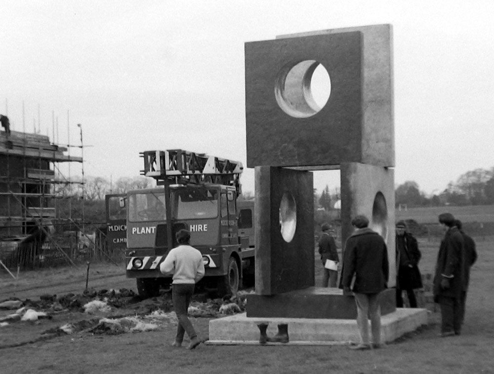 A black-and-white photo shows several people gathered near a large geometric sculpture with circular cutouts. A truck with Plant Hire on it is parked nearby. Scaffolding and a partially constructed building are visible in the background.