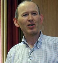 A person with short hair is speaking in front of a wooden background. They are wearing a checkered shirt.