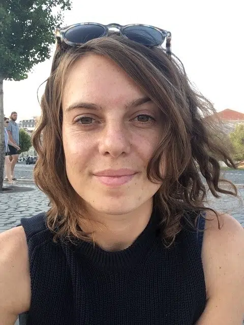 A person with wavy brown hair and sunglasses on their head smiles at the camera. She is wearing a sleeveless black top. In the background, there is a person walking on a stone path and some trees.