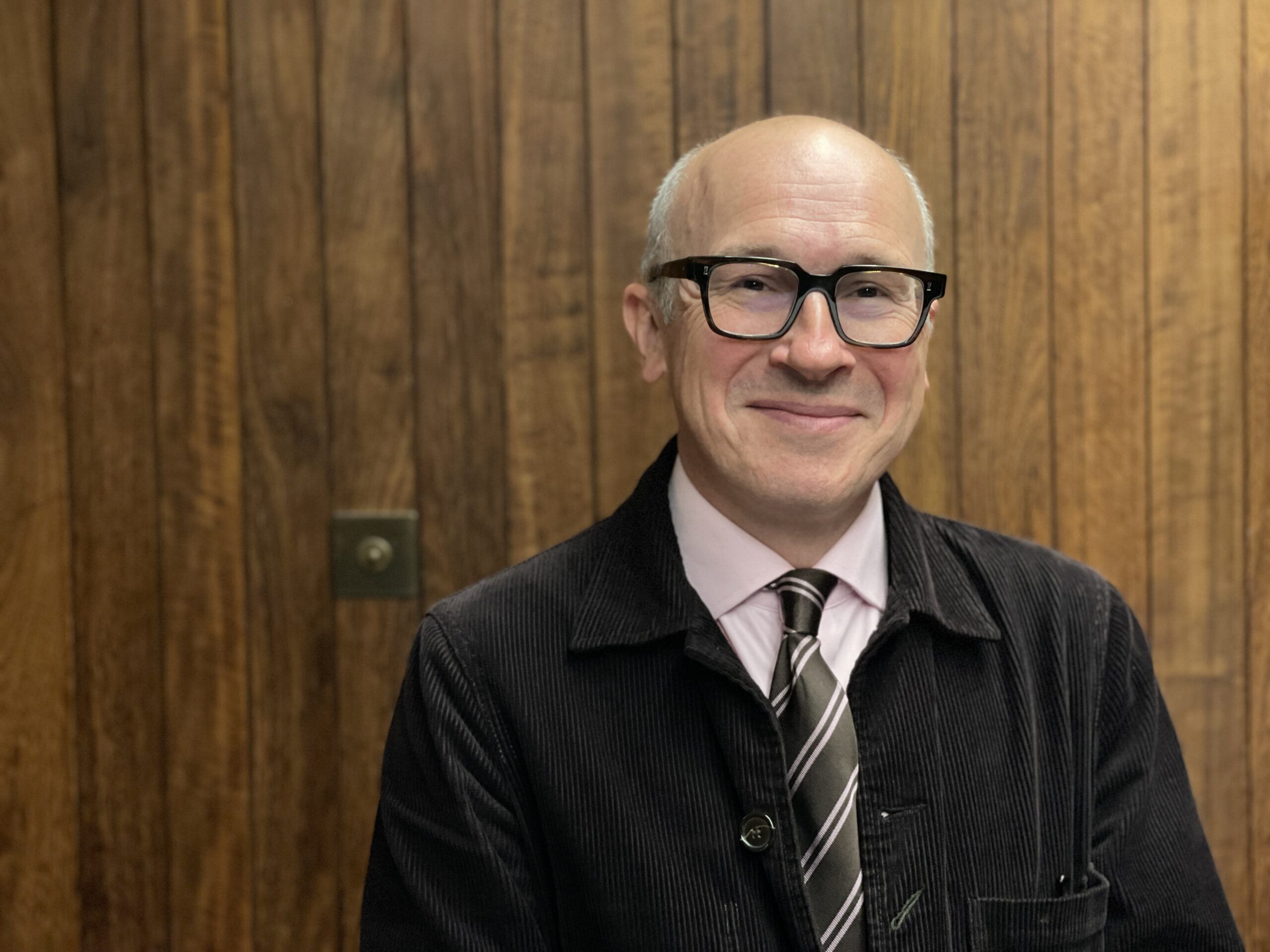 A bald man with glasses, wearing a black jacket, light pink shirt, and striped tie, stands in front of a wooden wall. He is smiling and looking directly at the camera.