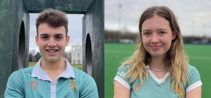 Two people are wearing light teal athletic jerseys with gold emblems. The person on the left is standing near a large metallic structure, while the person on the right is in front of a sports field. Both are smiling at the camera.