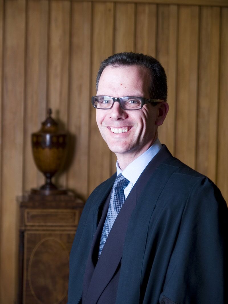 A person wearing glasses, a suit, and a dark robe smiles while standing in front of a wooden paneled wall. There is an ornate urn on a wooden cabinet beside them.