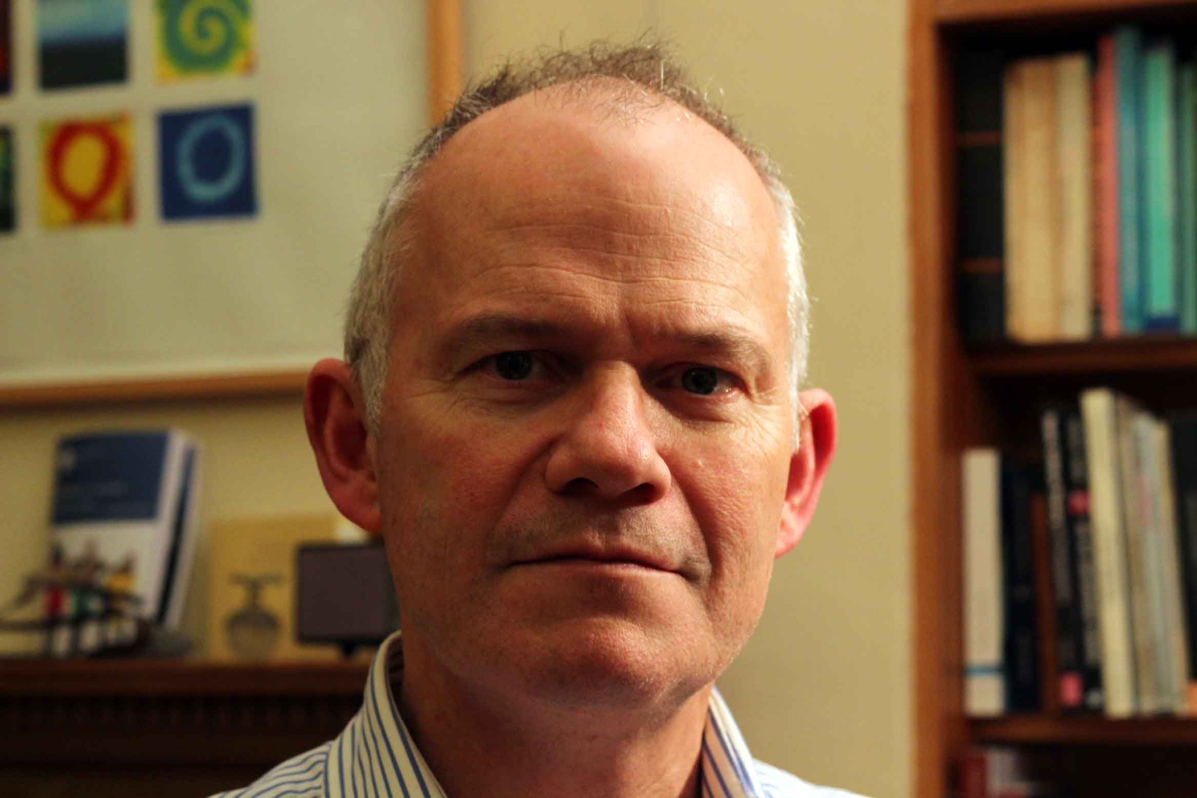 A man with a serious expression sits in a room with bookshelves and framed abstract art behind them. The person has short hair, and is wearing a striped shirt. The room has warm lighting.