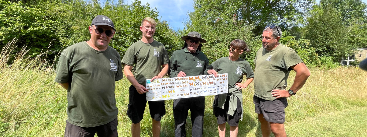 Five people in green shirts stand outdoors in a grassy area. They hold a poster with various butterfly images. Trees and bushes are visible in the background under a blue sky.