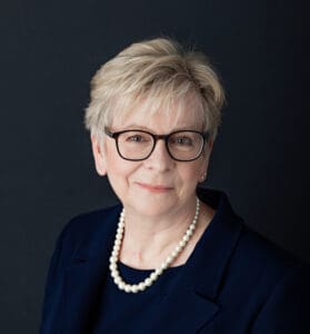Professor Sharon Peacock smiles at the camera. She has short light hair and glasses, and is wearing a pearl necklace and a dark blazer, against a dark background.