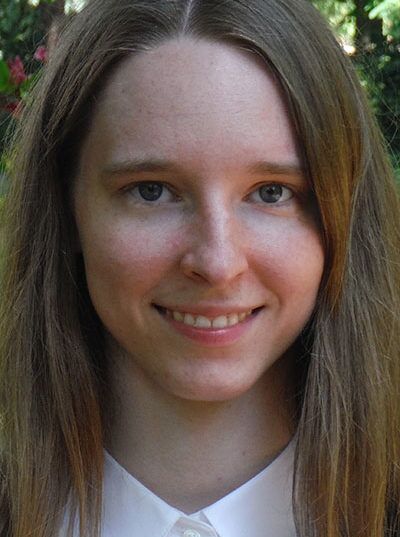 A person with long brown hair and light skin smiling directly at the camera. She is wearing a white collared shirt. The background is softly blurred greenery.