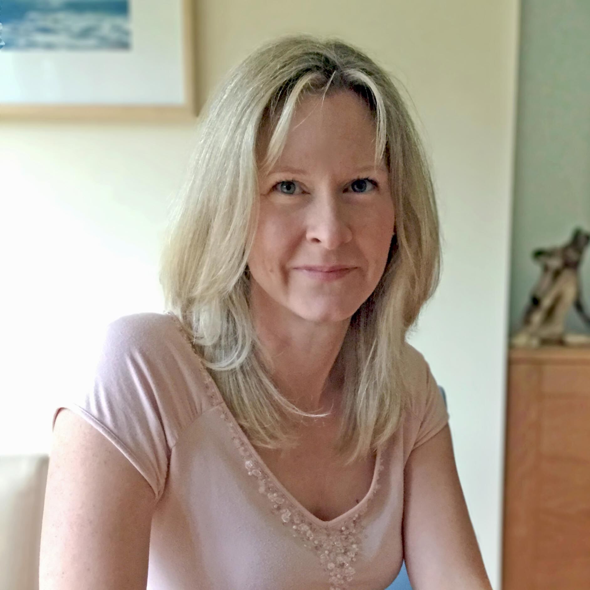 A woman with long blonde hair is smiling softly at the camera. She is wearing a light pink top and is indoors, with a blurred background featuring a wooden cabinet and a framed picture on the wall.